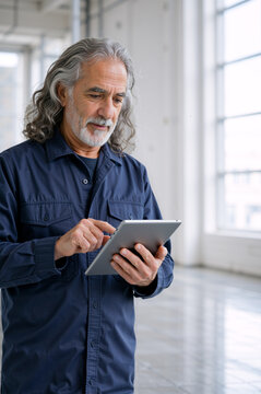 Senior man using digital tablet in bright empty industrial space. Vertical portrait of older male professional with gray hair working online. Copy space for text