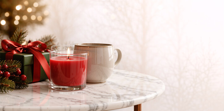 Lit red candle and ceramic mug next to green Christmas gift on marble table. Festive winter holiday composition with pine branches and bokeh lights. Copy space for text