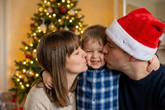 Happy mother and father wearing Santa hat kissing their young son near Christmas tree