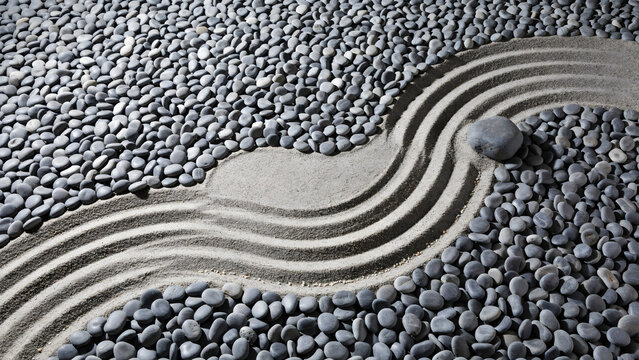 Zen garden scene with a single dark stone in a circular sand ripple pattern and flowing curved lines raked into the sand among grey pebbles
