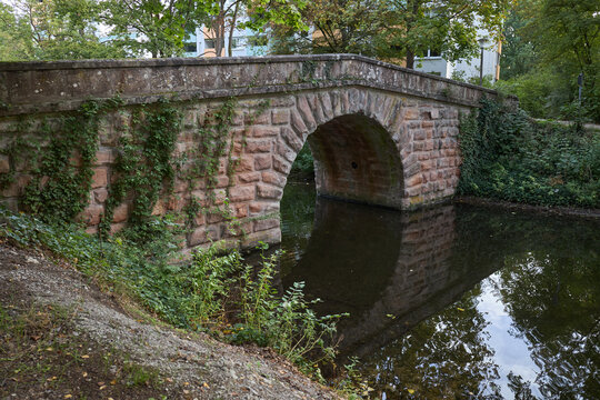 An ancient stone arch bridge crossing a calm stream in a lush green park setting in summer