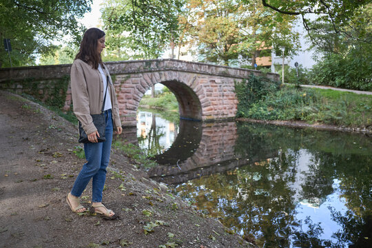 A woman standing on a path beside a tranquil canal with an old stone bridge in the background