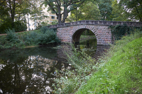 An old stone arch bridge spanning a calm canal surrounded by lush green trees and park foliage