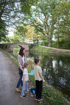 A mother and her two children standing by a canal and watching ducks in a peaceful park setting