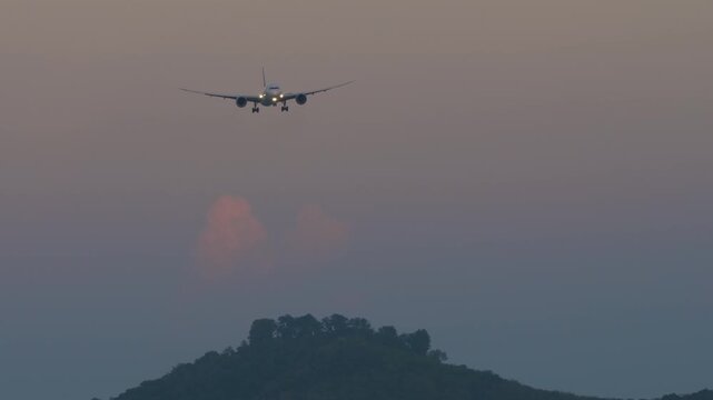 Commercial passenger airplane approaching for landing with landing lights on at sunset over forested green hills against twilight evening sky. Telephoto front view.