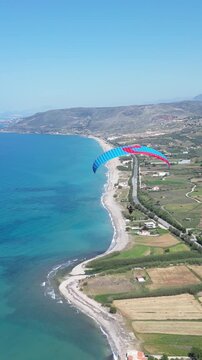 Vertical aerial view of paraglider flying along scenic Mediterranean sea coast with sandy beach and green fields on Crete island in Greece during summer. High quality FullHD footage