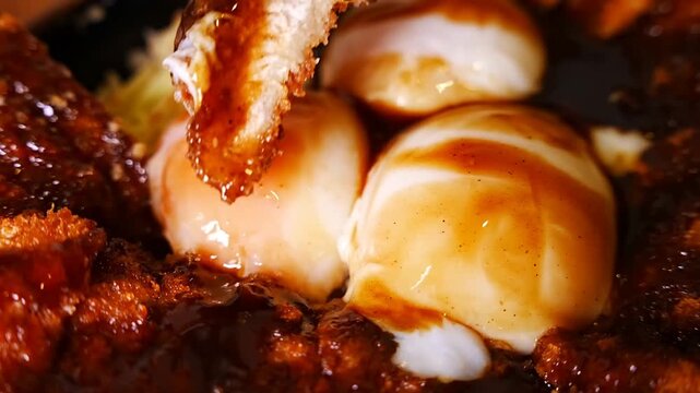 Close up view of a person using chopsticks to dip a piece of fried pork cutlet into a runny egg yolk inside a bowl filled with rice and dark sauce