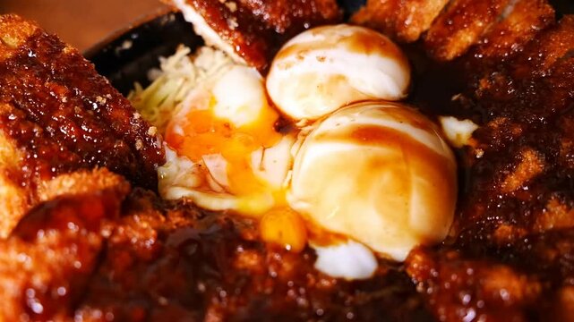 Close up view of a person using chopsticks to dip a piece of fried pork cutlet into a runny egg yolk inside a bowl filled with rice and dark sauce