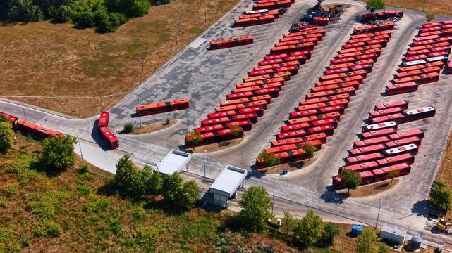 Multiple red buses stand in rows outdoors on sunny day. One coach is heading to the route. Aerial view.