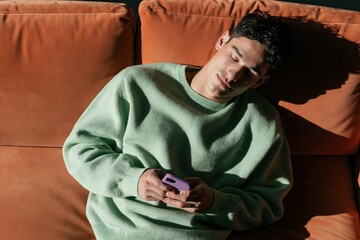 Fototapeta premium Young man asleep on sofa holding smartphone in sunlight for relaxed indoor background and lifestyle imagery