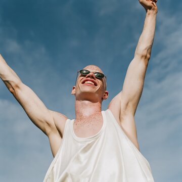Happy young man stretching arms upward wearing sunglasses under clear blue sky summer outdoor portrait expressing freedom and elation