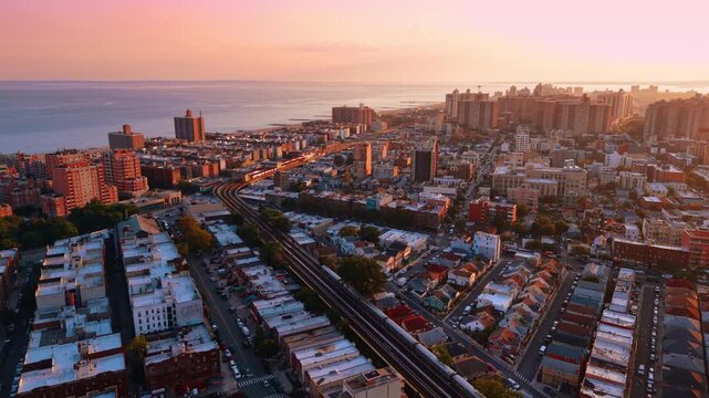Following a long train moving quickly by the railways across the cityscape. Scenery of New York City, USA against pink sky at sunset.