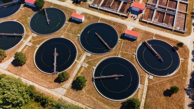 Big round reservoirs with dark water. Drone footage over the modern waste water treatment plant. Flock of birds flies over the facility.