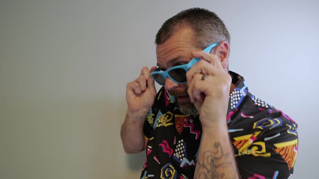 Middle aged man in 1980s graphic pattern shirt neon sunglasses and fanny pack posing humorously trying to look cool on neutral studio background