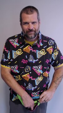 Middle aged man in 1980s graphic pattern shirt neon sunglasses and fanny pack posing humorously trying to look cool on neutral studio background
