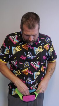 Middle aged man in 1980s graphic pattern shirt neon sunglasses and fanny pack posing humorously trying to look cool on neutral studio background