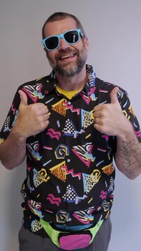 Middle aged man in 1980s graphic pattern shirt neon sunglasses and fanny pack posing humorously trying to look cool on neutral studio background