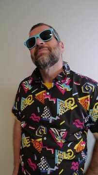Middle aged man in 1980s graphic pattern shirt neon sunglasses and fanny pack posing humorously trying to look cool on neutral studio background