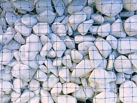 A top view of white smooth pebbles inside a metallic wire mesh gabion, forming a modern textured background used in landscaping, construction, and architectural design.