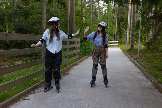 Two women on rollerblades raise their palms for a greeting on a forest skating track in summer