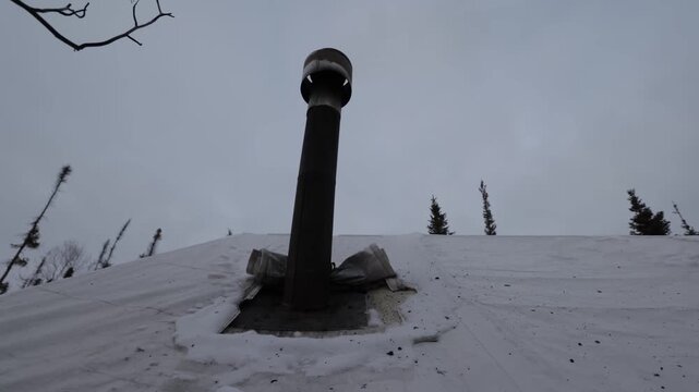 Low angle vent pipe rising from snow-covered roof in desolate alpine landscape under overcast sky, isolated windswept setting with distant evergreen horizon and debris