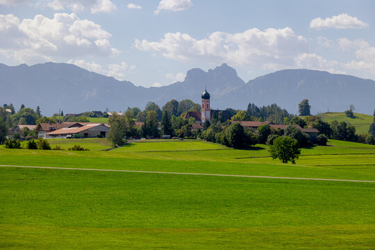 Countryside landscape, Traditional houses and church on rolling hill, Bavarian village with lush green grass meadow against majestic Alpine mountains under blue sky and white cloud in Bavaria, Germany