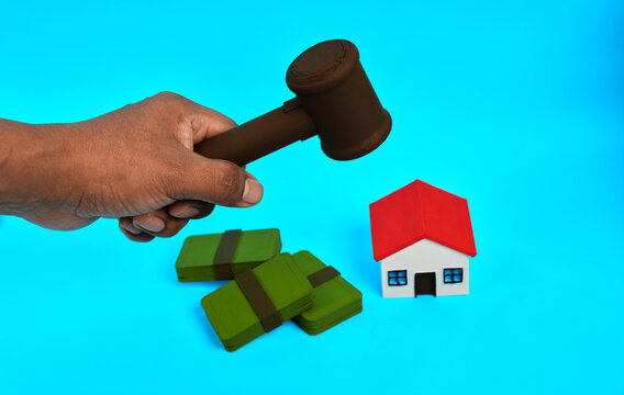 A hand holding a gavel above money bundles and a miniature house represents property auction and legal real estate transactions.
