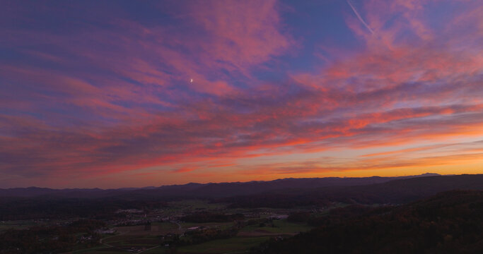 AERIAL: Fiery pink, purple and orange sunset spreads above forested hills and a quiet valley in Dolenjska, Slovenia. Autumn woodland and distant mountains create peaceful countryside panorama at dusk.