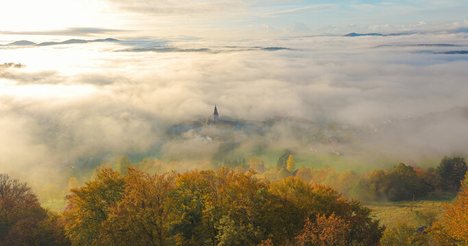 AERIAL: Sticna Abbey emerges softly through thick morning fog in peaceful autumn countryside of Slovenia. Golden trees frame the historic monastery while rolling hills rise above a vast sea of mist.