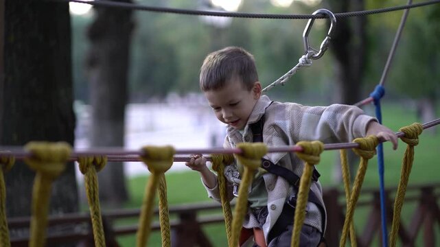 A young child explores a rope obstacle course in a safety harness, reaching forward with curiosity.