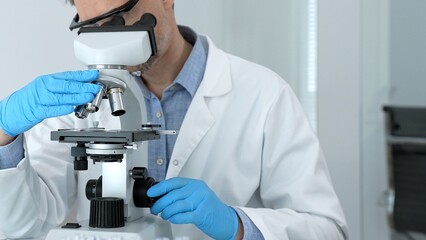 Scientist in a white lab coat and blue gloves examining samples using a modern microscope,...