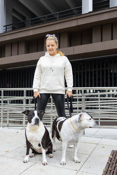 Young woman owner standing with her two pitbull walks dogs on leashes near modern city building