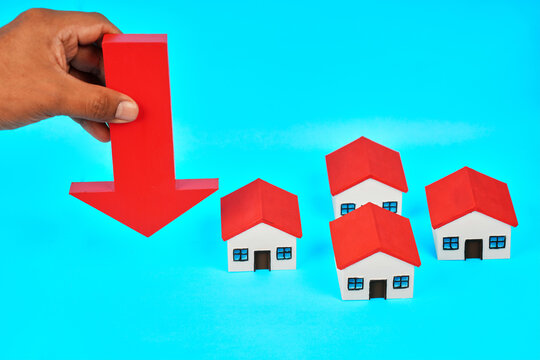A hand holding a downward arrow beside miniature houses represents falling property values and real estate market decline.
