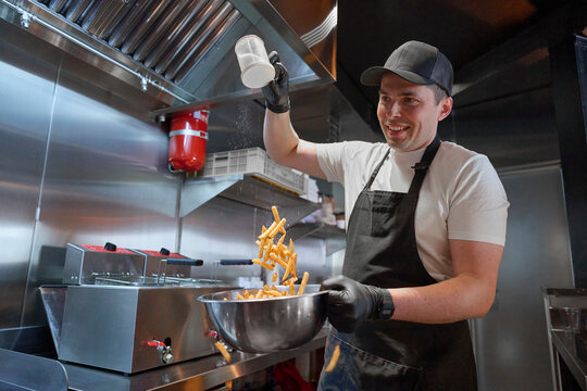 ook Adding Salt to Freshly Fried French Fries Just Taken from Fryer in Restaurant Kitchen