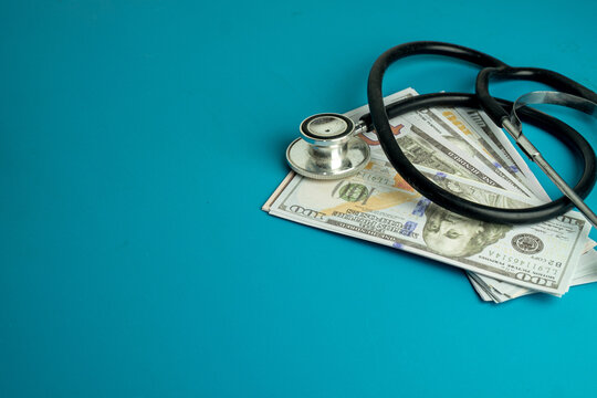 Black stethoscope resting on a stack of US dollar bills on a blue background with copy space. Concept of healthcare costs, medical insurance, expensive treatments and financial health.