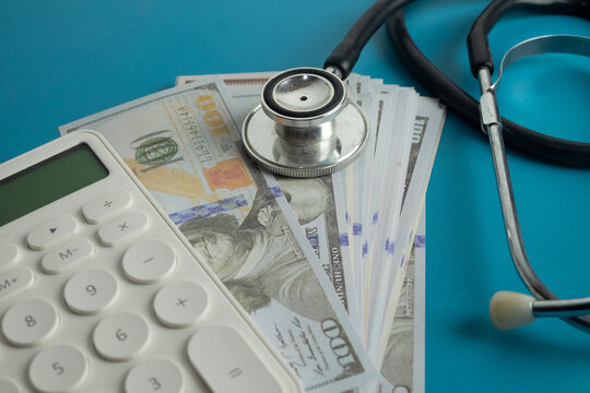 Stethoscope and white calculator on a stack of US dollar bills over blue background. Concept of calculating expensive healthcare costs, medical insurance, hospital fees and financial health.
