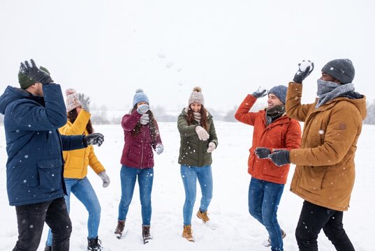 Group of happy friends having playful snowball fight in snowy field. Young men and women in colorful winter jackets, knitted hats and gloves throw snowballs during snowfall.