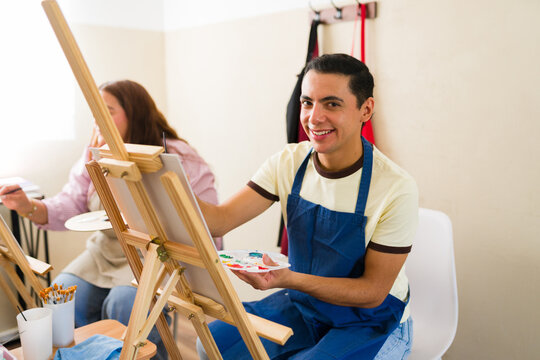 Smiling man enjoying an art class, painting on an easel while holding a color palette, exploring his creativity