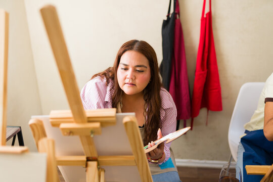 Woman concentrating during painting class, holding palette and creating artwork on canvas easel in art studio