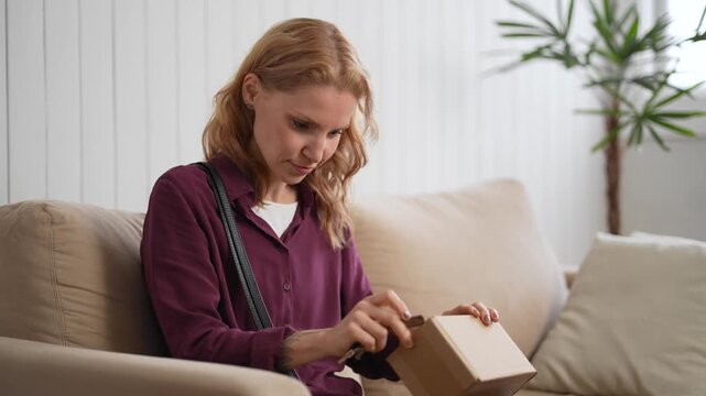 Excited woman sitting on the sofa, lifting a small white box from an open parcel and examining it closely. Happy female customer checking a new online shopping purchase