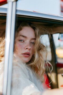 Close-up lifestyle portrait of a young blonde woman sitting inside a car, framed by an open window in bright natural daylight