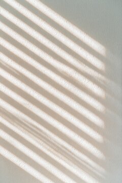 Minimal beige wall with diagonal sunlight and soft window blind shadows, creating a warm neutral abstract background with copy space