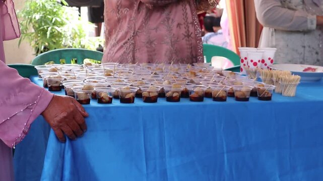 Traditional Indonesian party catering table with rows of savory snacks served in plastic cups with toothpicks and sweet spicy sauce.