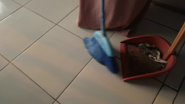 A person sweeping a ceramic tile floor with a broom while using a dustpan to collect dirt and dust. The household cleaning activity represents routine housework, floor maintenance, hygiene, and keepin