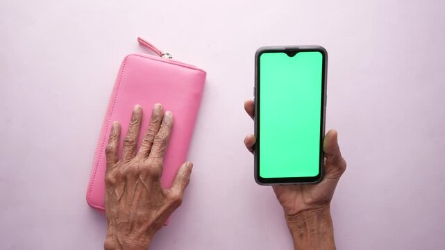 Elderly woman hands with wrinkles holding a green screen smartphone over a pink background with a pink purse