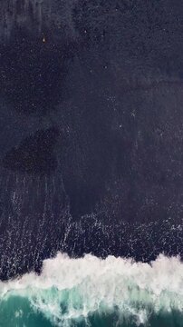 Aerial looking down at waves on a black sand beach in Bali Indonesia. Vertical Video.