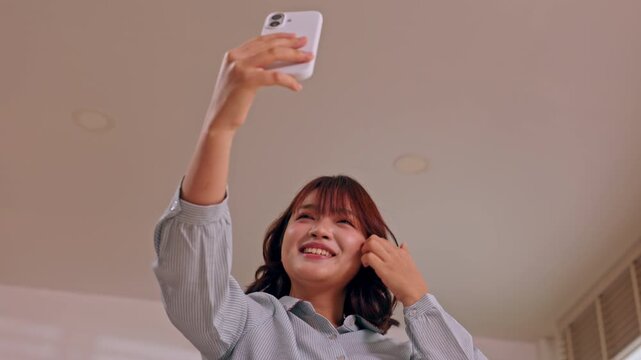 Asian woman holding smartphone with raised arm, smiling indoors during casual selfie at home, holding headphone with cheerful expression, modern lifestyle moment viewed from below