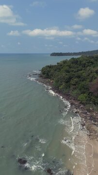 Vertical backward aerial view shows a lush, forested headland meeting the rocky, windy coastline, where waves wash over the beach under a bright blue, partly cloudy sky.