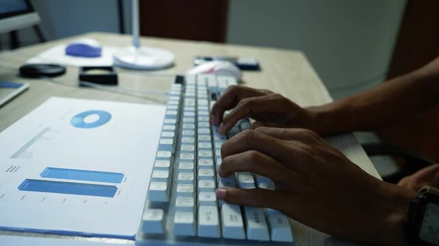 Hands typing on a white computer keyboard beside printed business charts on an office desk, showing data analysis and workplace productivity.
