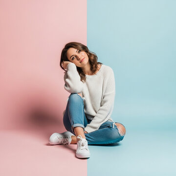 Young Woman Sitting on Floor in Ripped Jeans and Sweater Against Pink and Blue Background